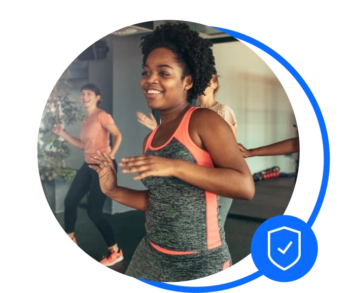 Zumba instructor with a black afro is dancing in a Zumba class. She's wearing a grey gym set with a coral stripe down the side and waving her hands to the side and smiling ahead.